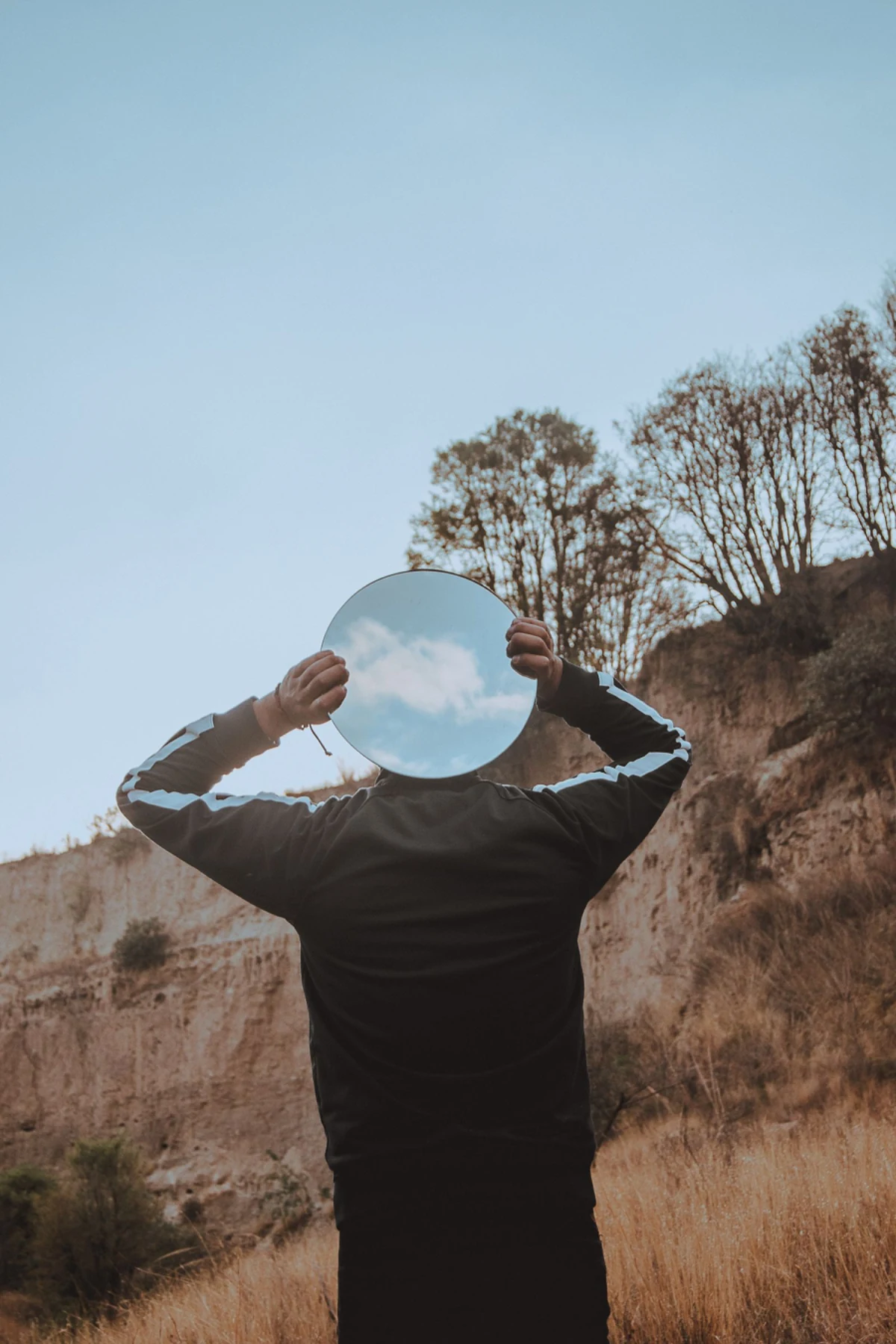 A person holds a circular mirror over their face, reflecting blue sky in a dry hillside landscape.