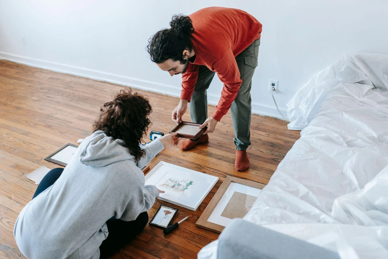 A couple looks through framed pictures beside a covered sofa during a move.